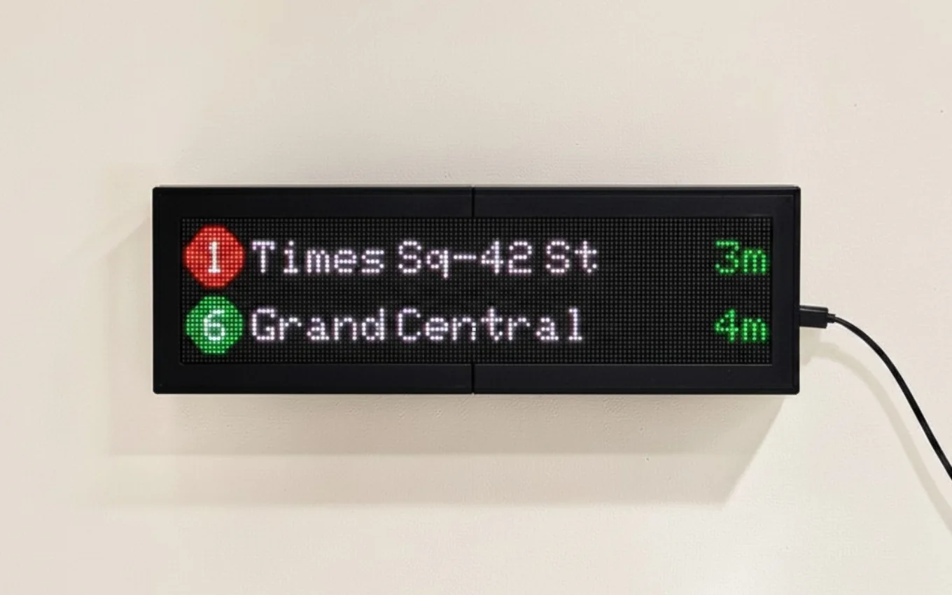 A CommuteLive LED board mounted on a plain cream wall, showing arrivals for Times Sq-42 St on the 1 train in 3 minutes and Grand Central on the 6 train in 4 minutes